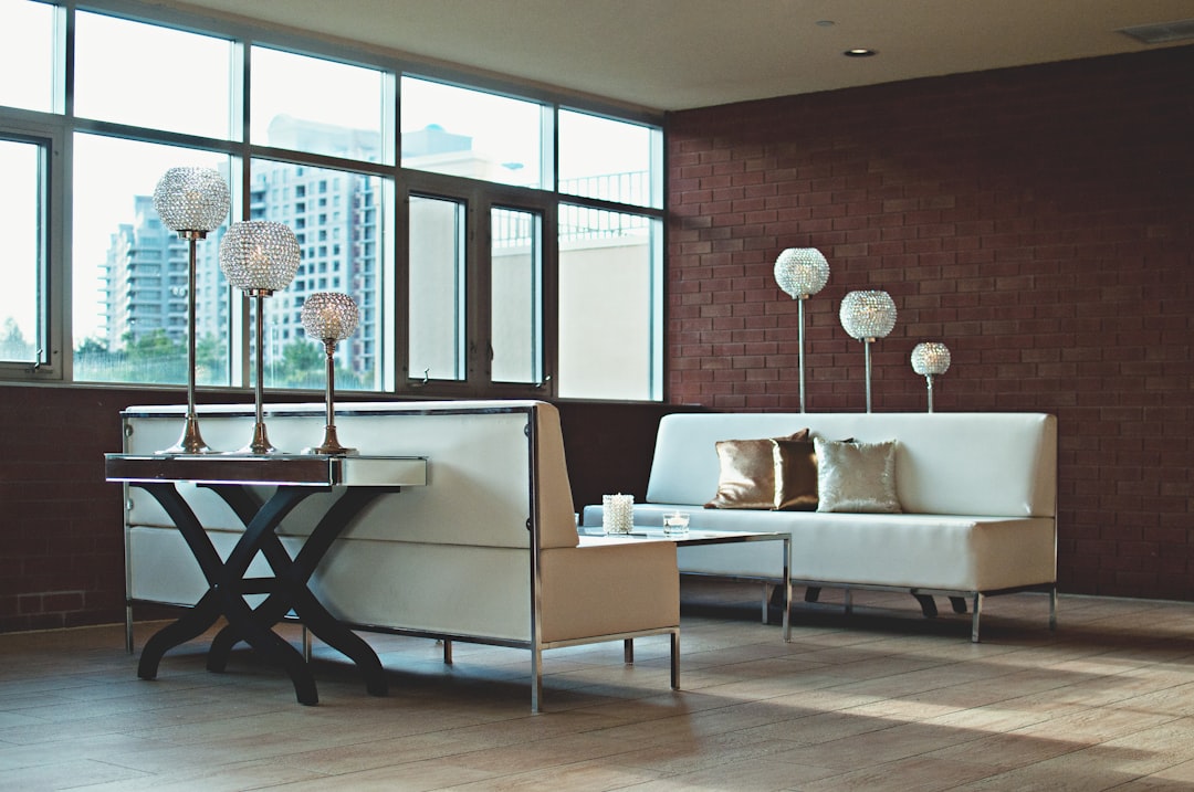 Modern living room with white leather sofas, crystal floor lamps, and a glass coffee table set against a red brick wall and large windows overlooking city buildings.