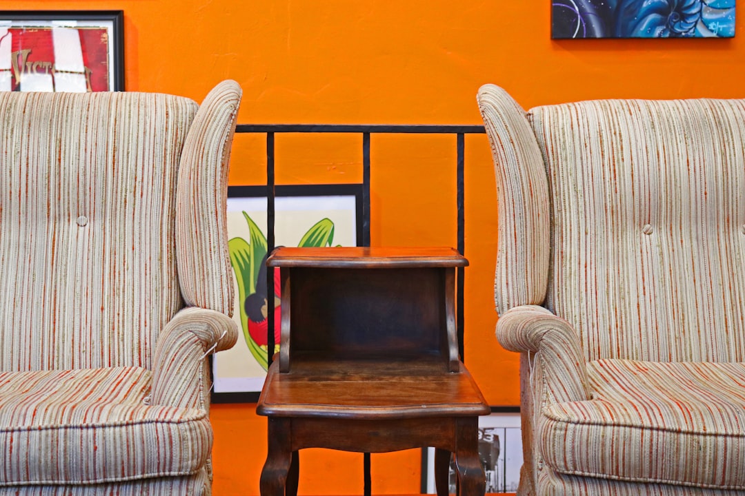 Two vintage striped armchairs with a wooden side table between them against a bright orange wall with framed artwork.