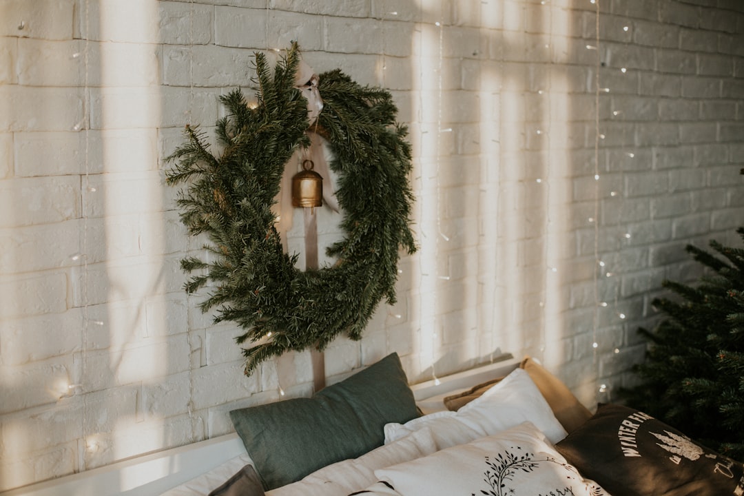 A large green Christmas wreath with a hanging bell displayed on a white brick wall above cozy pillows, with soft holiday lights creating a warm, festive atmosphere.