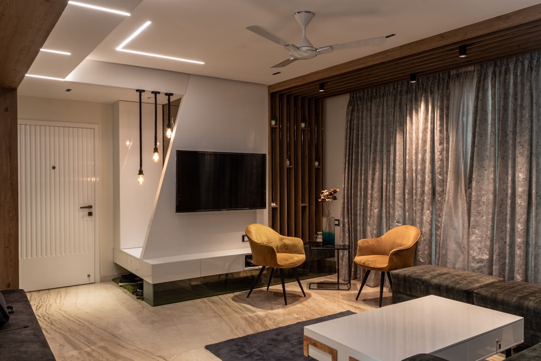 A stylish living room with a geometric TV wall, strip lighting, marble floor, and two mustard yellow accent chairs facing a dark sofa