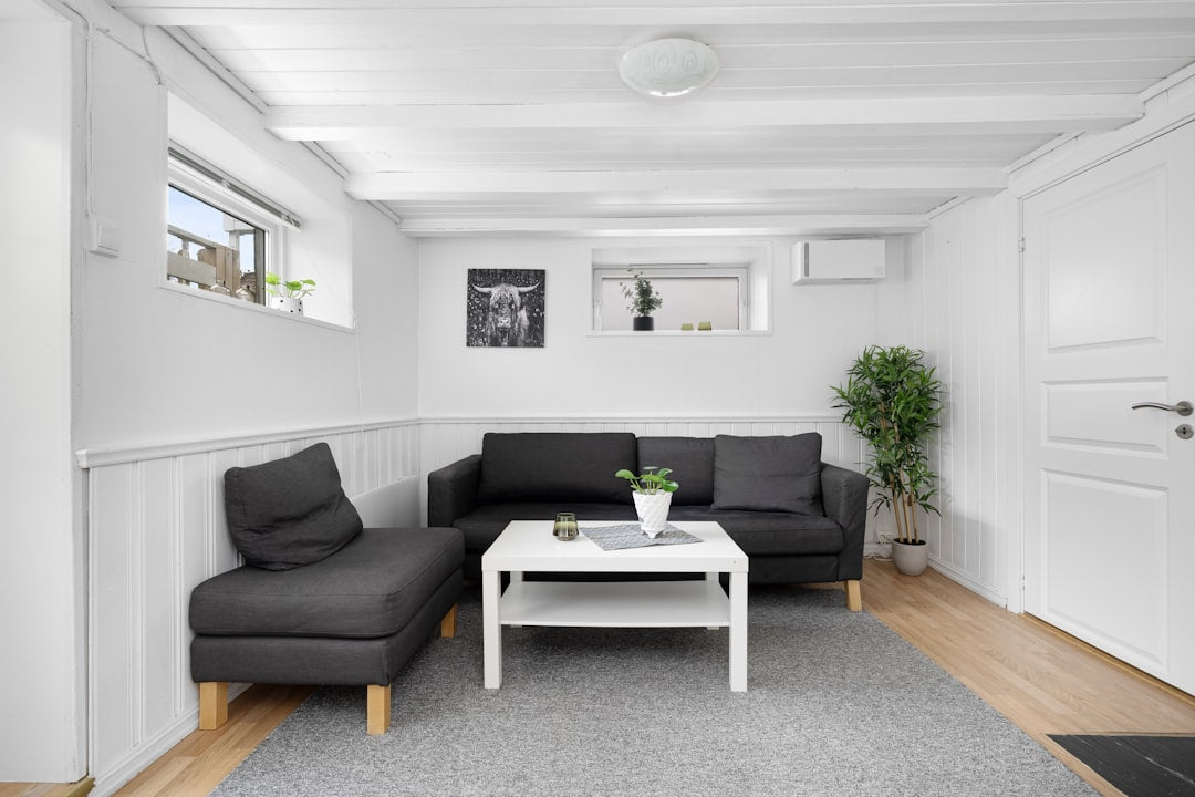 Minimal white basement living room with dark gray sectional sofa, white coffee table, indoor plants, small windows and light wood flooring.