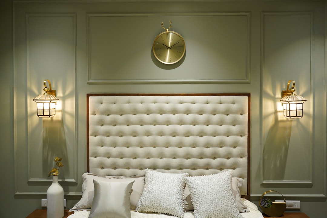 A symmetrical shot of a luxurious bedroom with pale green wainscoting and molding, featuring a large tufted, off-white upholstered headboard, a brass clock mounted above it, and two antique-style brass lantern wall sconces on either side.